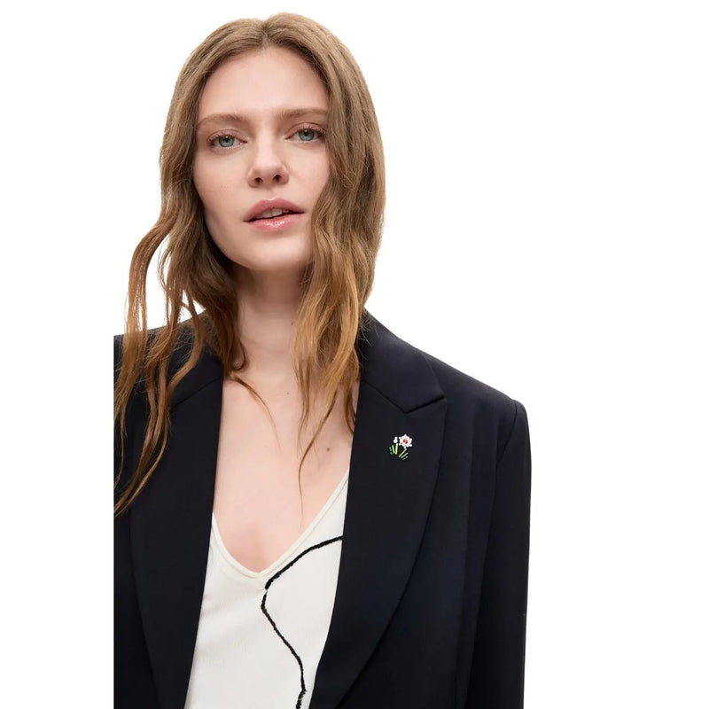 Eine zarte, florale Metallbrosche mit weißer Blüte und grünem Stiel an einem schwarzen Desigual-Damenblazer aus Viskosemischung mit langem Revers