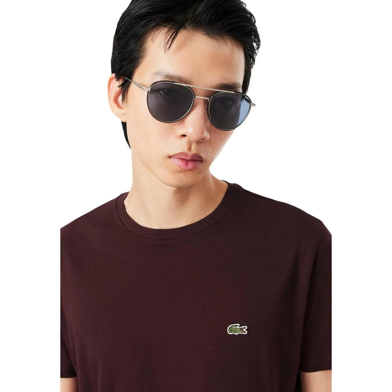 Silberfarbene Pilotenbrille mit dunkelgrauen Gläsern, kombiniert mit einem bordeauxroten Baumwoll-T-Shirt von Lacoste für Herren.