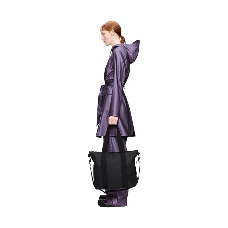Eine elegante schwarze Mini-Tragetasche von Rains für Damen aus glänzendem Polyester mit stabilen Henkeln, gehalten von einem Model in einem violetten Regenmantel.
